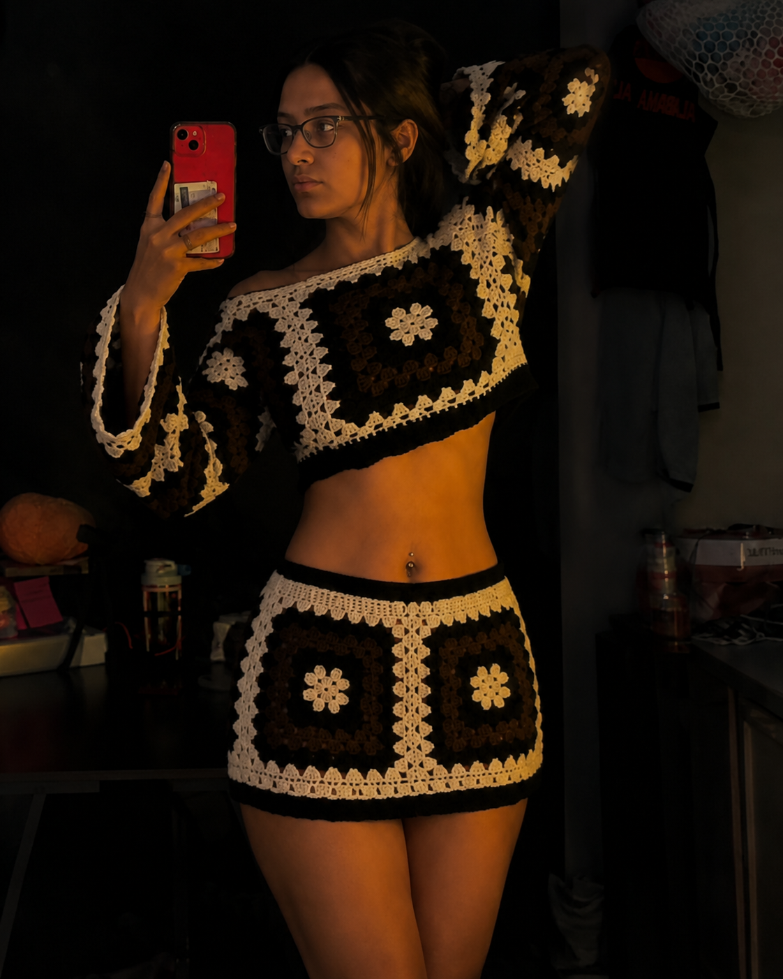 A woman takes a mirror selfie wearing a matching crochet two-piece set consisting of a cropped long-sleeve top and a mini skirt. The crochet pattern features alternating dark brown and cream/off-white squares with floral motifs.